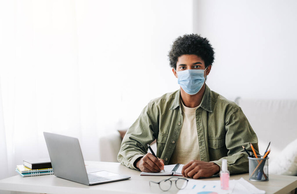 Edukacja internetowa podczas covid-19. czarny nastolatek facet w chirurgiczne maska robi notatki podczas online wykład na laptop - Zdjęcie, obraz