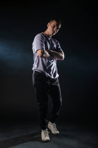 Handsome young man wearing a gray t-shirt and black pants poses on black background in a studio - Fotó, kép