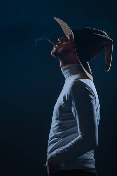 Портрет красивого молодого человека в ковбойской шляпе, курящего сигарету в студии на высоком контрасте и темном фоне - Фото, изображение
