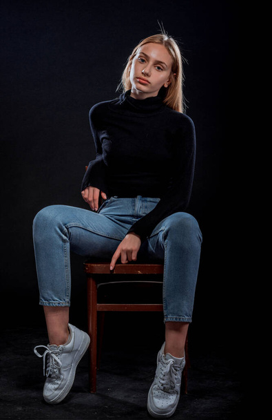 Πορτρέτο ενός όμορφου κομψού κοριτσιού που φοράει μαύρο πουλόβερ και τζιν κάθεται σε ξύλινο cheir - Φωτογραφία, εικόνα