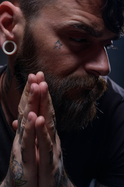 Közelkép egy szakállas férfiról tetoválással a kezén, arcán és nyakán, fekete ingben.. - Fotó, kép