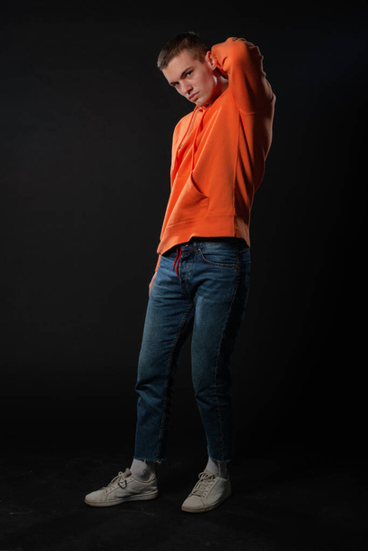 Молодой красивый мужчина в джинсах и оранжевой толстовке позирует, стоя на черном фоне. - Фото, изображение