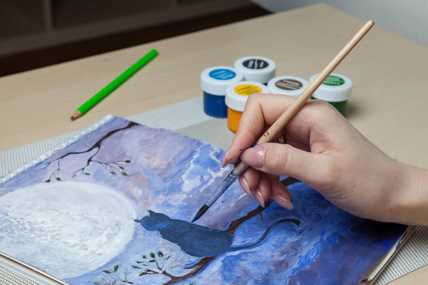 As mãos femininas mantêm uma escova e uma cobertura de um jarro de aquarela e misturam pinturas contra o contexto de um gato em uma árvore, o céu noturno e a lua perto de ferramentas de trabalho na mesa. Belas artes em modo de quarentena. - Foto, Imagem