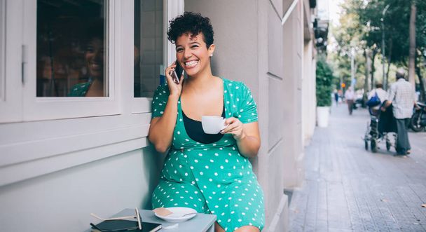Smiling bright ethnic lady in casual wear using mobile talking to friend relaxing drinking cup of coffee at outside of cafe - Photo, image