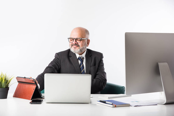 Asiatico indiano anziano uomo d'affari finanziario seduto alla sua postazione di lavoro o scrivania di fronte a un computer, laptop e tablet. Parlare al telefono mentre si fa qualche scartoffia - Foto, immagini