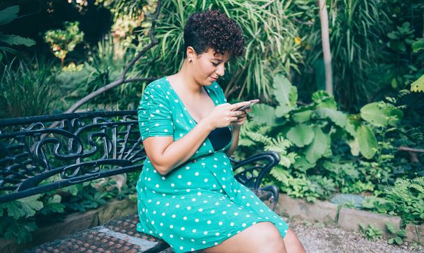From above of plump African American woman in casual green gown watching video on mobile phone while sitting on bench at summer park - Фото, зображення