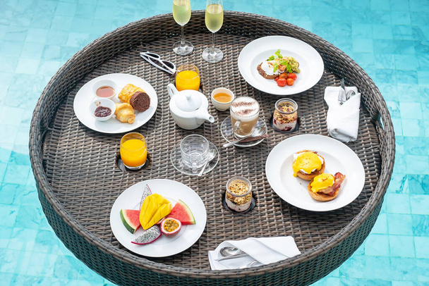 Pływająca taca śniadaniowa w basenie w luksusowym hotelu lub tropikalnej willi kurortu, owoce; mango, smok arbuz i marakuja owoce, jedzenie, chleb, kawa, herbata, wino i sok pomarańczowy. Egzotyczne lato - Zdjęcie, obraz