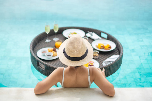Szczęśliwa kobieta w białym stroju kąpielowym o pływające śniadanie taca w luksusowym hotelu basen, młoda kobieta z kapeluszem cieszyć się w tropikalnym ośrodku. Relaks, egzotyczne podróże letnie, wakacje, wakacje i weekend - Zdjęcie, obraz