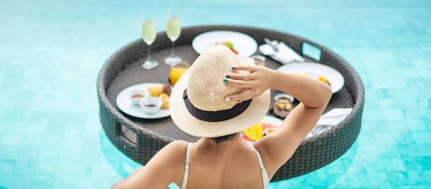 高級プールホテルでフローティングブレックファーストトレイを持っている白い水着の幸せな女性、帽子を持つ若い女性は熱帯リゾートでお楽しみください。リラックス、エキゾチックな夏の旅行、休日、休暇、週末 - 写真・画像