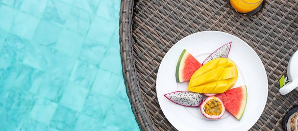 Pływająca taca śniadaniowa w basenie w luksusowym hotelu lub tropikalnej willi kurortu, owoce; mango, owoce smoka arbuza, owoce pasji i sok pomarańczowy. Egzotyczne podróże letnie i koncepcja zdrowej żywności - Zdjęcie, obraz