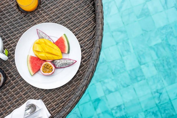 Floating Breakfast tray in swimming pool at luxury hotel or tropical resort villa, fruits; mango, watermelon dragon fruit, passion fruit and orange juice. Exotic summer travel and healthy food concept - Photo, Image