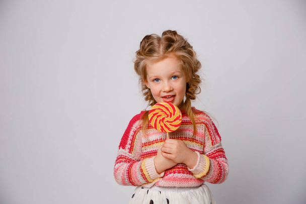 retrato de una niña una niña rubia con una piruleta en un palo sonriendo con ropa de color sobre un fondo blanco - Foto, imagen