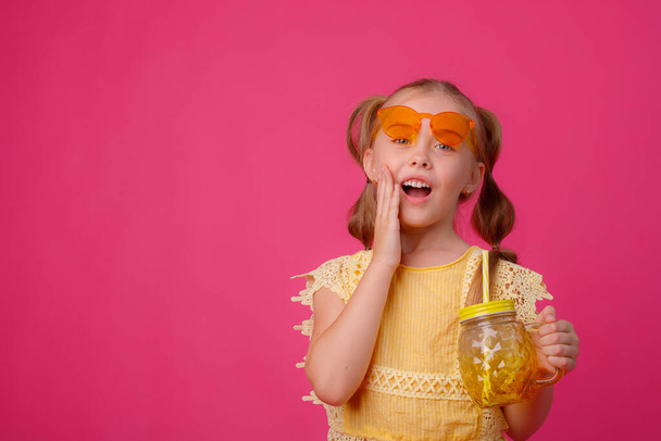 una niña pequeña en gafas de sol con un cóctel sobre un fondo rosa - Foto, Imagen