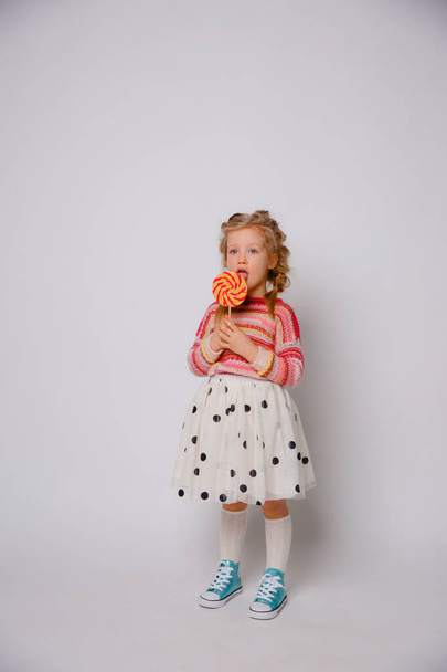 retrato de una niña una niña rubia con una piruleta en un palo sonriendo con ropa de color sobre un fondo blanco - Foto, Imagen