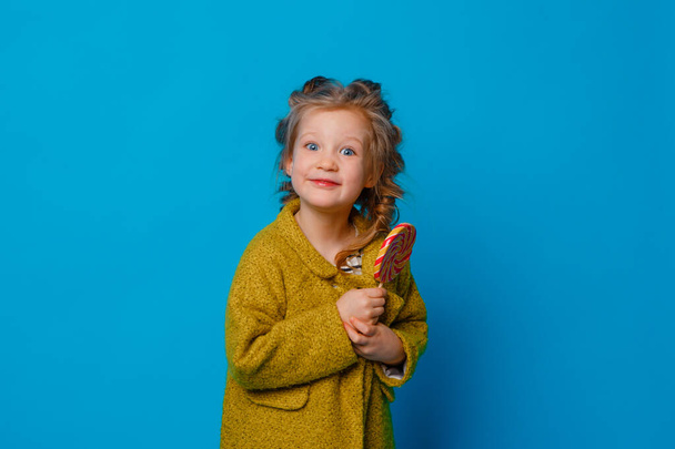 portrait of a baby girl in a coat holding a Lollipop on a colored blue background - Photo, Image