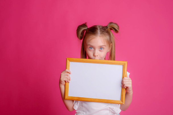 Portrait of a girl holding an empty frame, looks out for the text Studio on a pink background - Foto, imagen