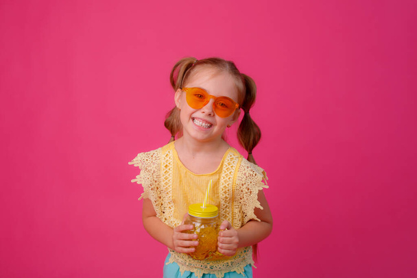 portré egy szőke kislányról napszemüvegben rózsaszínen egy üveg hűtő koktéllal. - Fotó, kép
