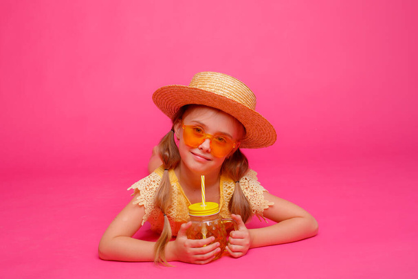 a little girl in sunglasses , a straw hat and a cocktail is lying on a pink background - Фото, зображення