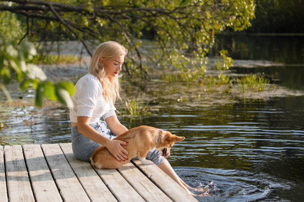 Safkan Galli Corgi Pembroke köpeği olan kadın gölün kıyısındaki ahşap iskelede oturuyor. - Fotoğraf, Görsel