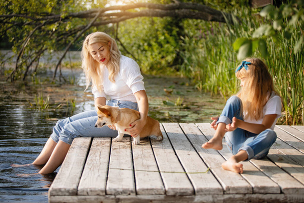 女性と彼女の娘と純血草のウェルシュコーギー刺繍犬は湖で木製の桟橋に座っています - 写真・画像