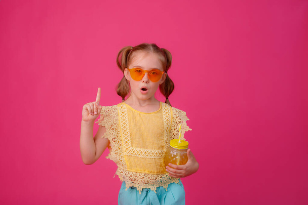 una niña pequeña en gafas de sol con un cóctel sobre un fondo rosa - Foto, Imagen