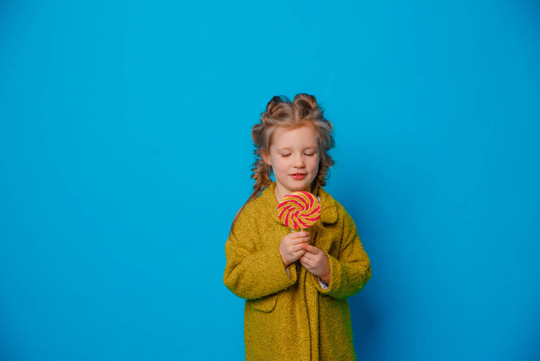Porträt eines kleinen Mädchens in einem Mantel, das einen Lutscher auf einem farbigen blauen Hintergrund hält - Foto, Bild