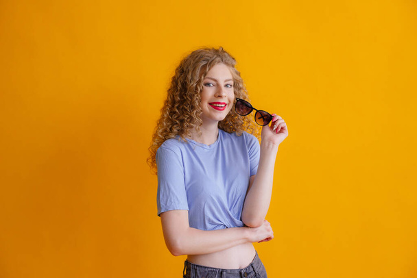 curly ginger woman posing with sunglasses against orange background - Foto, afbeelding