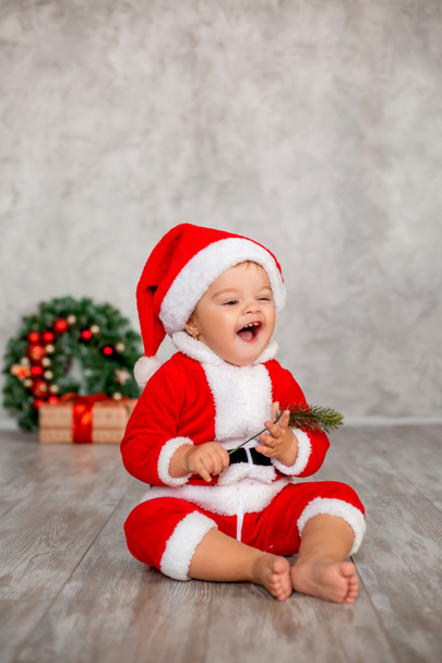 Sweet baby Santa sits on a wooden floor with a gift and a Christmas wreath. Christmas concept, text space - Photo, Image