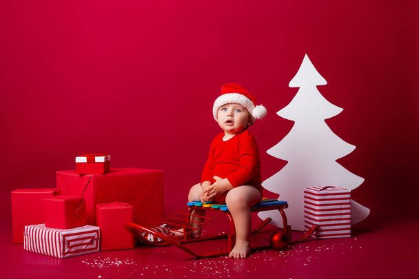 Cute baby Santa sits on a sled with Christmas gifts. Christmas concept, text space - Photo, Image