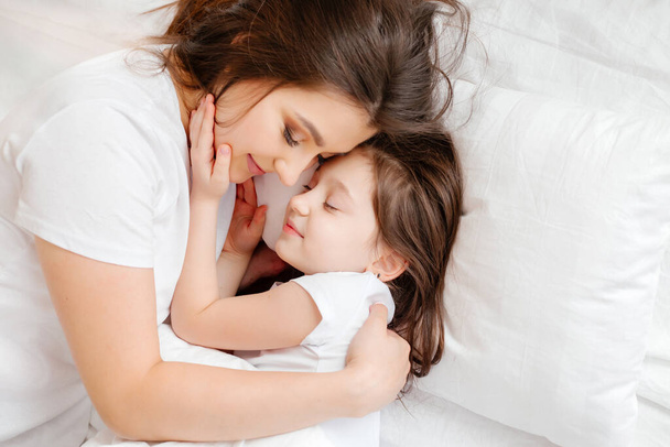 Šťastná mladá matka objímá svou dcerku v posteli, pohled shora. matka a dcera odpočívají doma v posteli. Dobré ráno! - Fotografie, Obrázek