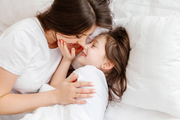 Onnellinen nuori äiti halaa pientä tytärtään sängyssä, näkymä ylhäältä. äiti ja tytär lepäävät sängyssä kotona. Huomenta.! - Valokuva, kuva