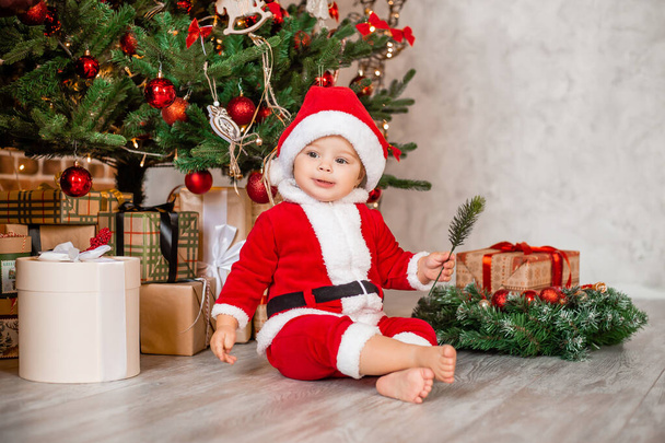 Симпатичный малыш Санта сидит дома возле елки с подарками - Фото, изображение