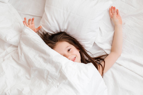 Ευτυχισμένο κοριτσάκι ξαπλώνει στο κρεβάτι στο σπίτι το πρωί. υγιή ύπνο του μωρού. λευκά κλινοσκεπάσματα, χώρος για κείμενο - Φωτογραφία, εικόνα