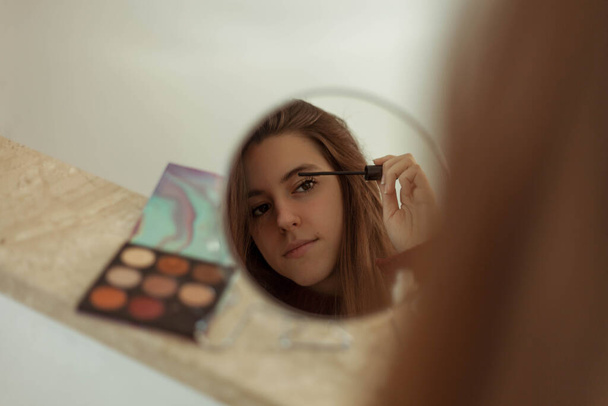 Όμορφη νεαρή γυναίκα που βάζει μακιγιάζ μπροστά από έναν καθρέφτη. Νεαρή καστανή γυναίκα - Φωτογραφία, εικόνα