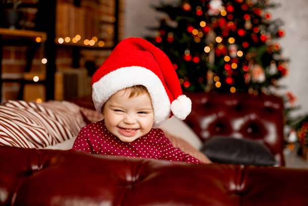 Cute baby Santa siedzi w domu w pobliżu choinki z prezentami - Zdjęcie, obraz