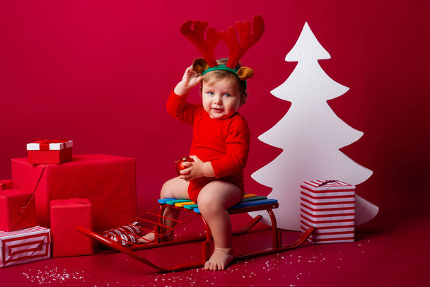 Cute kid with deer antlers on his head sits on a sledge with Christmas gifts. concept of kinship, space for text - Fotó, kép