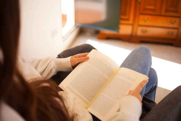 mujer joven leyendo un libro sentado en un sofá. Mujer con cabello castaño - Foto, imagen