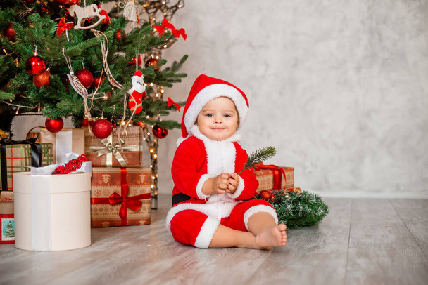 Niedliche Baby-Santa sitzt zu Hause neben dem Weihnachtsbaum mit Geschenken - Foto, Bild