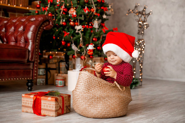 Cute baby Santa sits at home near the Christmas tree with gifts - Fotó, kép
