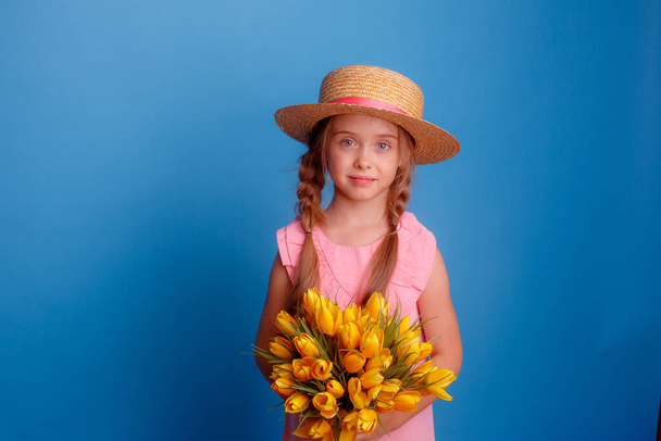 ein kleines Mädchen mit Strohhut hält einen Strauß gelber Tulpen auf blauem Hintergrund - Foto, Bild