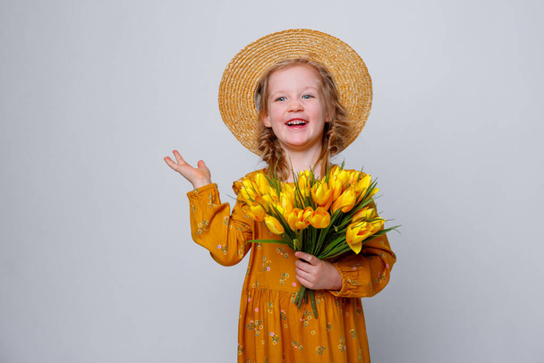 κοριτσάκι με ψάθινο καπέλο και ένα μπουκέτο ανοιξιάτικα λουλούδια, απομονωμένο σε λευκό φόντο. Κορίτσι παιδί με ένα μπουκέτο κίτρινες τουλίπες - Φωτογραφία, εικόνα