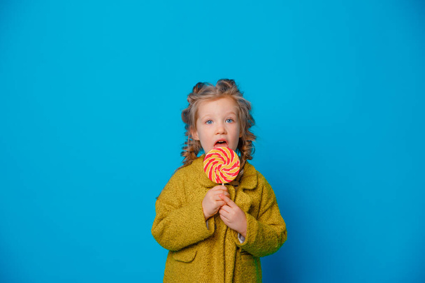 portrait of a baby girl in a coat holding a Lollipop on a colored blue background - Фото, зображення