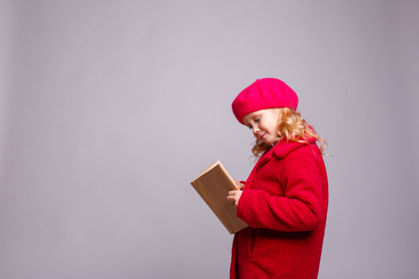 little schoolgirl girl in a coat and beret with a book  against  gray background - Photo, Image