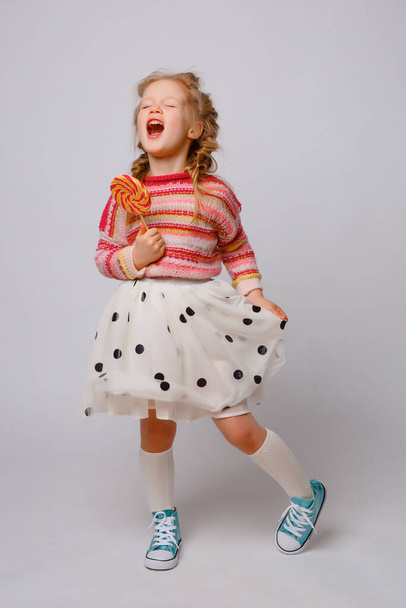 portret van een meisje een blond kind met een Lollipop op een stok glimlachen in gekleurde kleren op een witte achtergrond - Foto, afbeelding