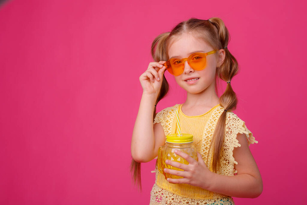 πορτρέτο ενός μικρού ξανθού κοριτσιού με γυαλιά ηλίου σε ροζ χρώμα με ένα γυάλινο βάζο δροσιστικό κοκτέιλ. - Φωτογραφία, εικόνα