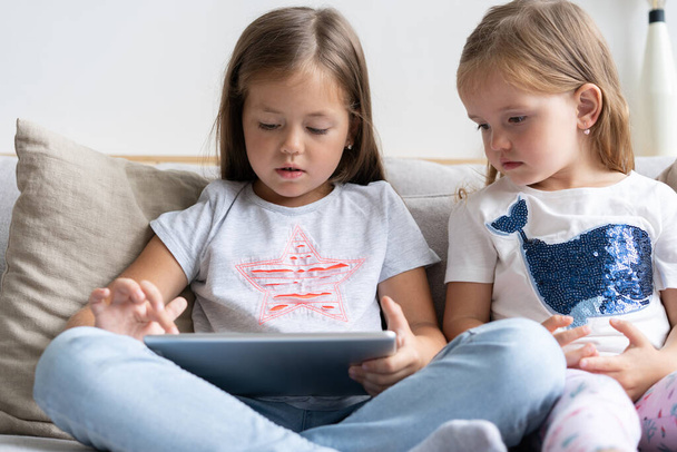 Little girls or sisrers sitting on the sofa playing digital tablet in the living room at home. - Фото, изображение