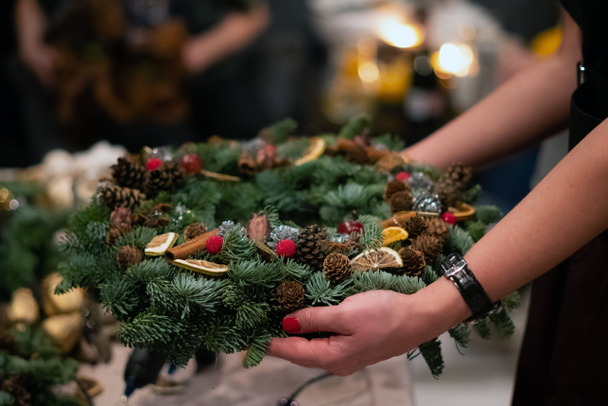 Χριστουγεννιάτικο εργαστήριο ύφανσης στεφανιών. Γυναικεία χέρια στολίζουν στεφάνι διακοπών από κλαδιά ερυθρελάτης, κώνους και διάφορες οργανικές διακοσμήσεις στο τραπέζι - Φωτογραφία, εικόνα