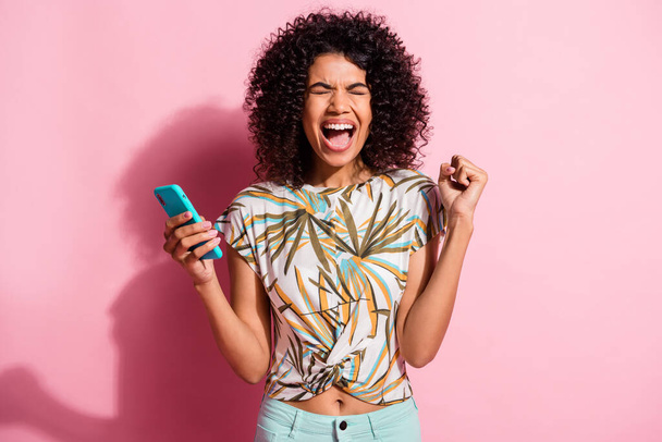 Photo portrait of screaming woman celebrating holding phone in one hand with fist in air isolated on pastel pink colored background - Foto, afbeelding