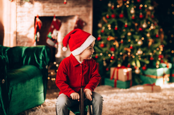 A fiú egy ló formájú fa hintán ül. Karácsonyi hangulat. A háttérben egy díszített karácsonyfa és egy nagy kandalló. - Fotó, kép