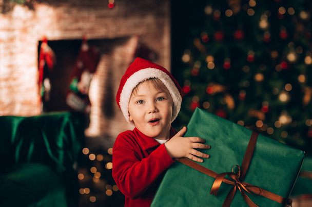 Um menino segurando uma caixa grande em um pacote verde e um arco com um presente. Humor de Natal. Contra o fundo de uma grande árvore de Natal e uma lareira. - Foto, Imagem